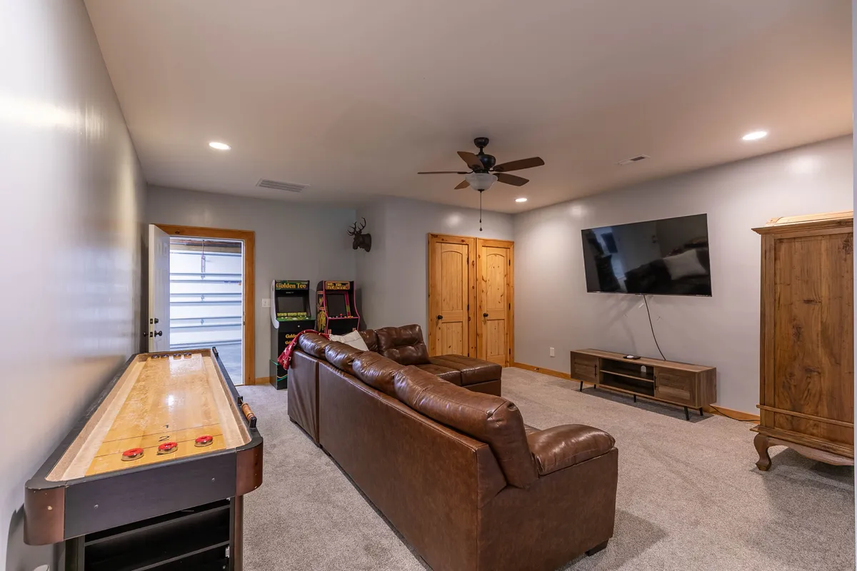 A casual, inviting family game room featuring a leather sofa, television, and a shuffleboard table, all set against a backdrop of warm wood and soft lighting. The room is arranged for both relaxation and interactive fun, with the shuffleboard table positioned for easy play and the TV setup calling out for movie nights.