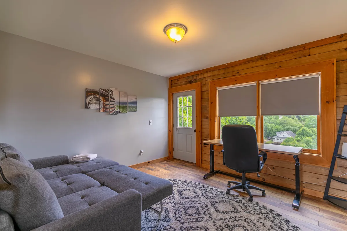 A cozy upstairs loft with a comfortable sofa, desk, large windows, and a warm wooden wall. The space blends relaxation with potential for games or quiet activities, illuminated by natural light. The atmosphere is intimate and inviting, ideal for board games or kids’ hangouts.