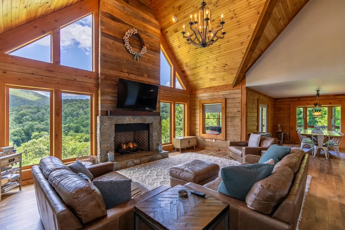 A cozy living room scene with a stacked-stone fireplace as the focal point, surrounded by plush leather sofas, a rustic chandelier above, and large windows framing beautiful sunset views over the mountains and farmland. The mood is warm and inviting for gatherings and quiet moments alike.