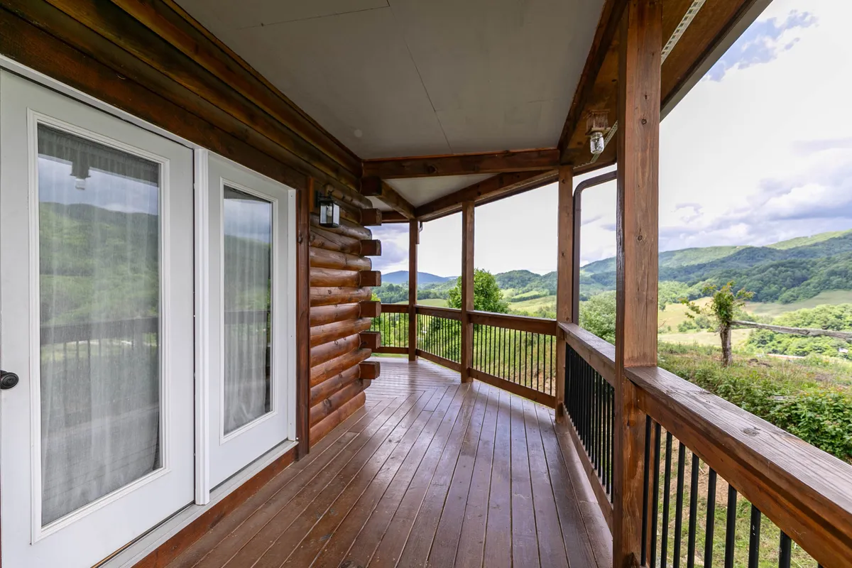 A log cabin scene with a wide porch, wooden railing, double doors, and panoramic views of surrounding mountains and forests. The porch is perfect for gatherings or quiet moments, offering an inviting spot to enjoy coffee in the morning or relax in the evening light.