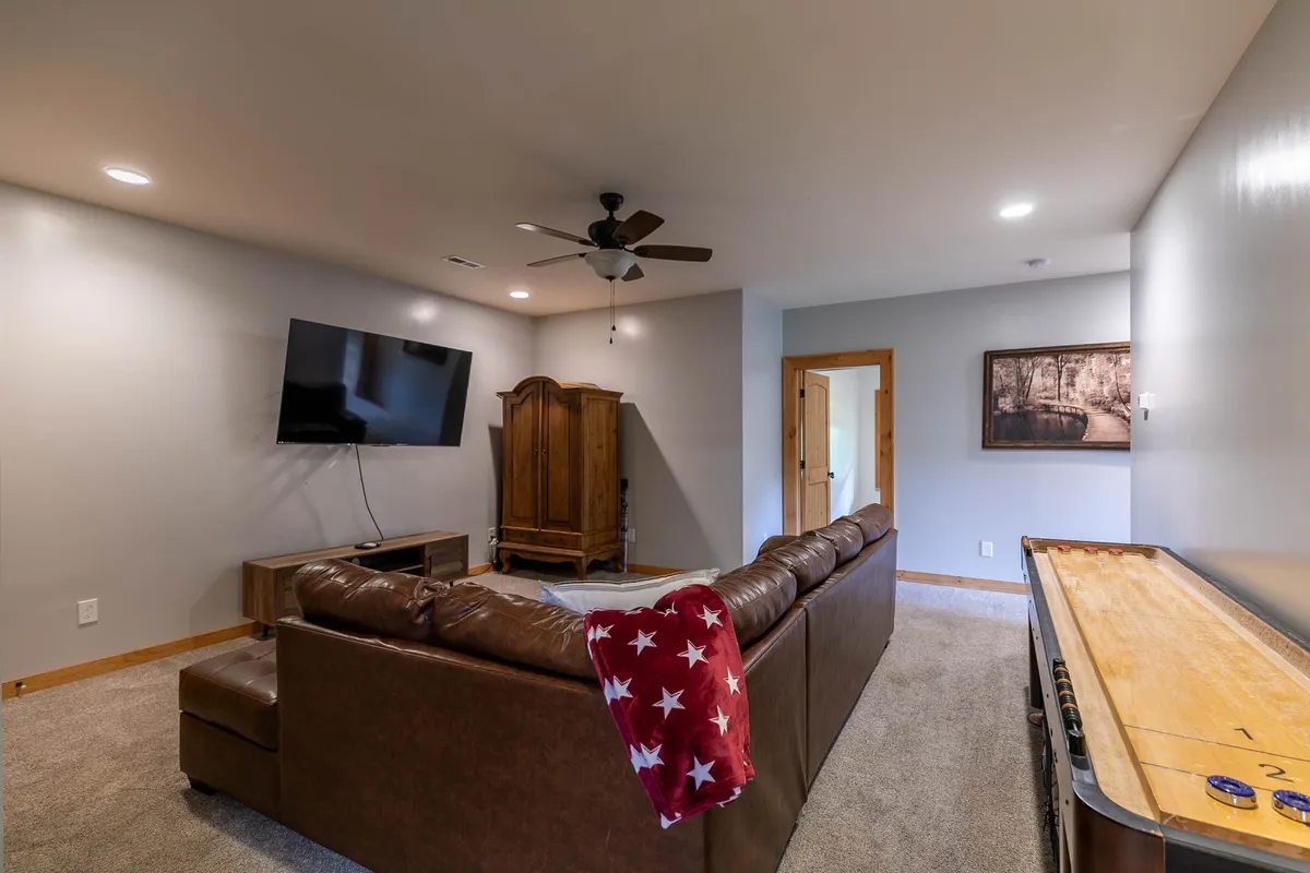 A spacious, relaxed game room scene with a large brown leather sectional couch, a wall-mounted TV, a prominent shuffleboard table, and tasteful decorative elements. The arrangement encourages group gatherings and friendly competition, with ample space for lounging and engaging in games.