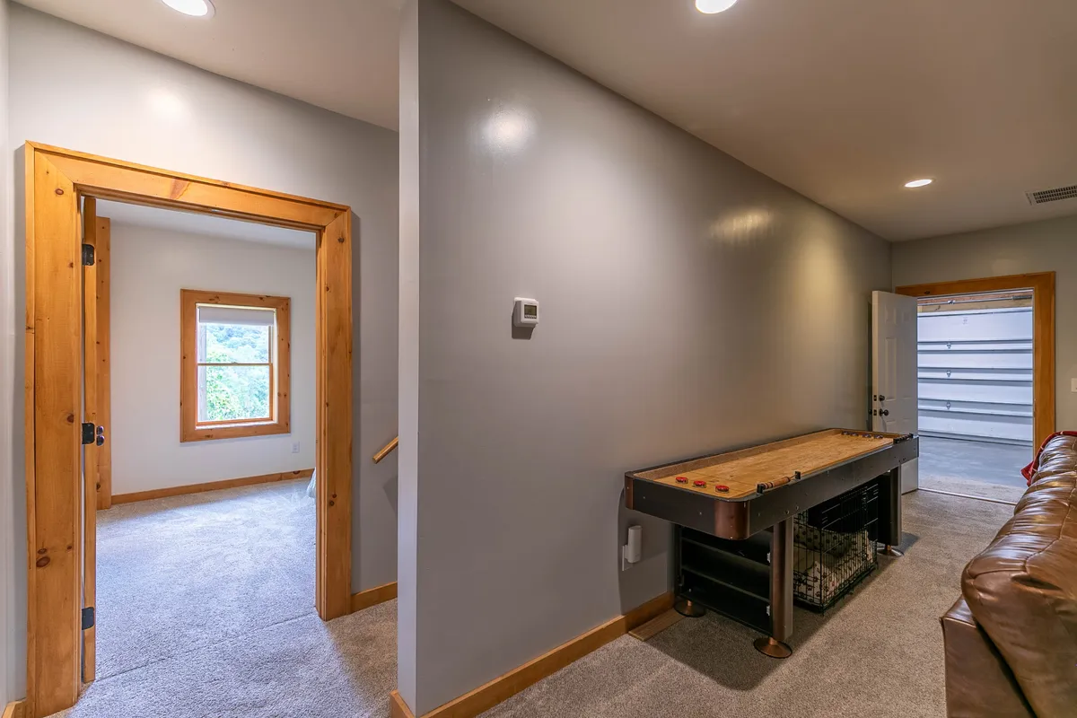 A casual, welcoming game room with a shuffleboard table, open doorway, and a glimpse of the garage door. The space invites socializing, friendly competition, and comfortable downtime for all ages.