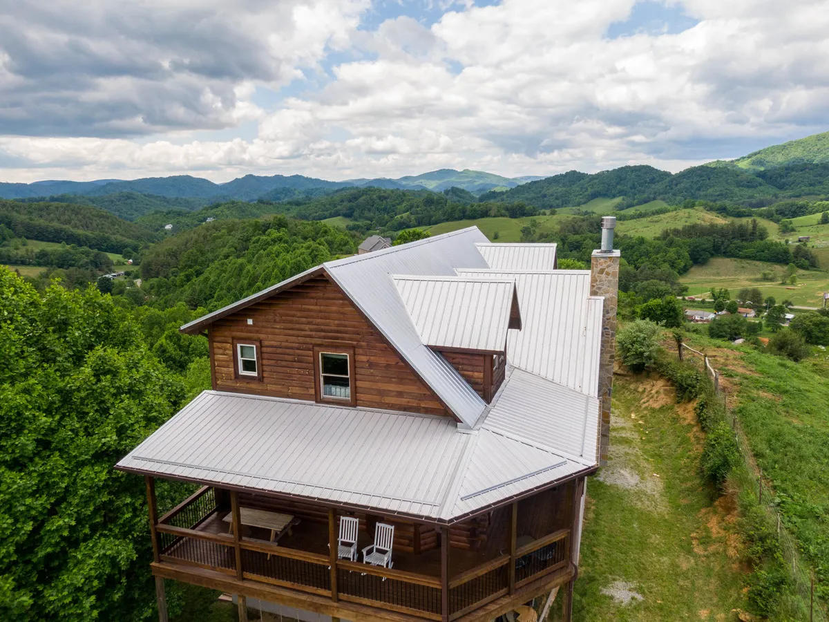 A charming wooden cabin with a metal roof sits against a backdrop of mountains. The porch is furnished with chairs, inviting guests to relax and soak in the tranquil mountain setting. Lush greenery surrounds the cabin, and the open porch connects directly to nature, suggesting easy access for outdoor walks and adventures.