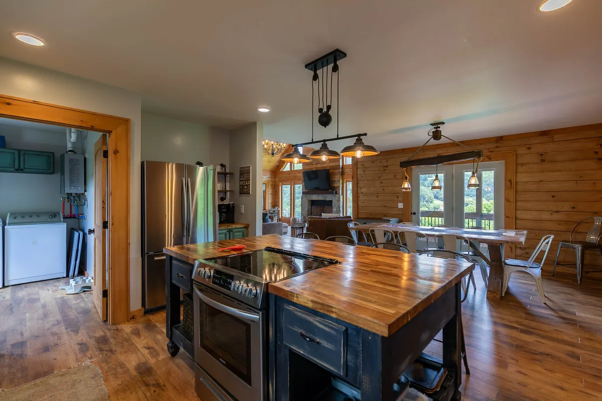 A bright, open kitchen and dining area featuring a spacious kitchen island with barstools, stainless steel appliances, a wooden dining table, and large windows that let in ample natural light and offer scenic views. The layout encourages social cooking and shared meals.