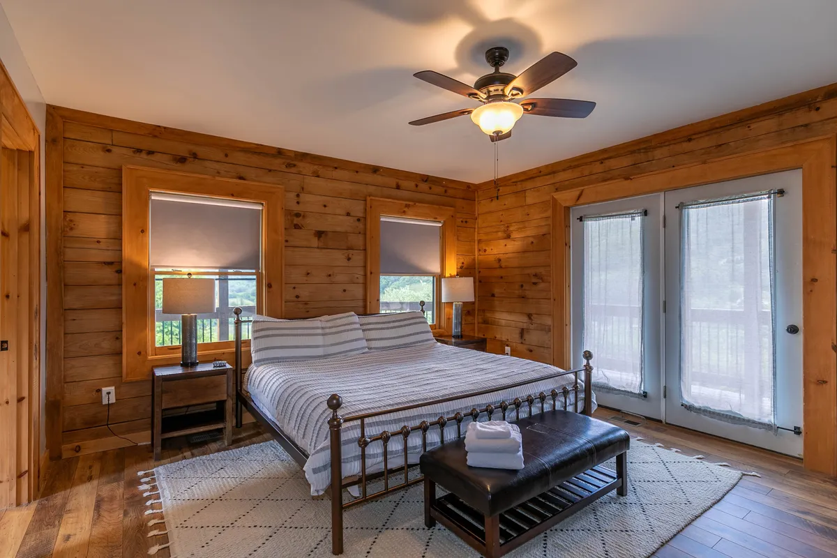 A tranquil bedroom scene featuring a cozy bed with striped bedding, warm wooden walls, a nightstand with a lamp, and double doors that open directly to the outdoors. Natural light streams into the room, and there is a clear sense of peace and relaxation.