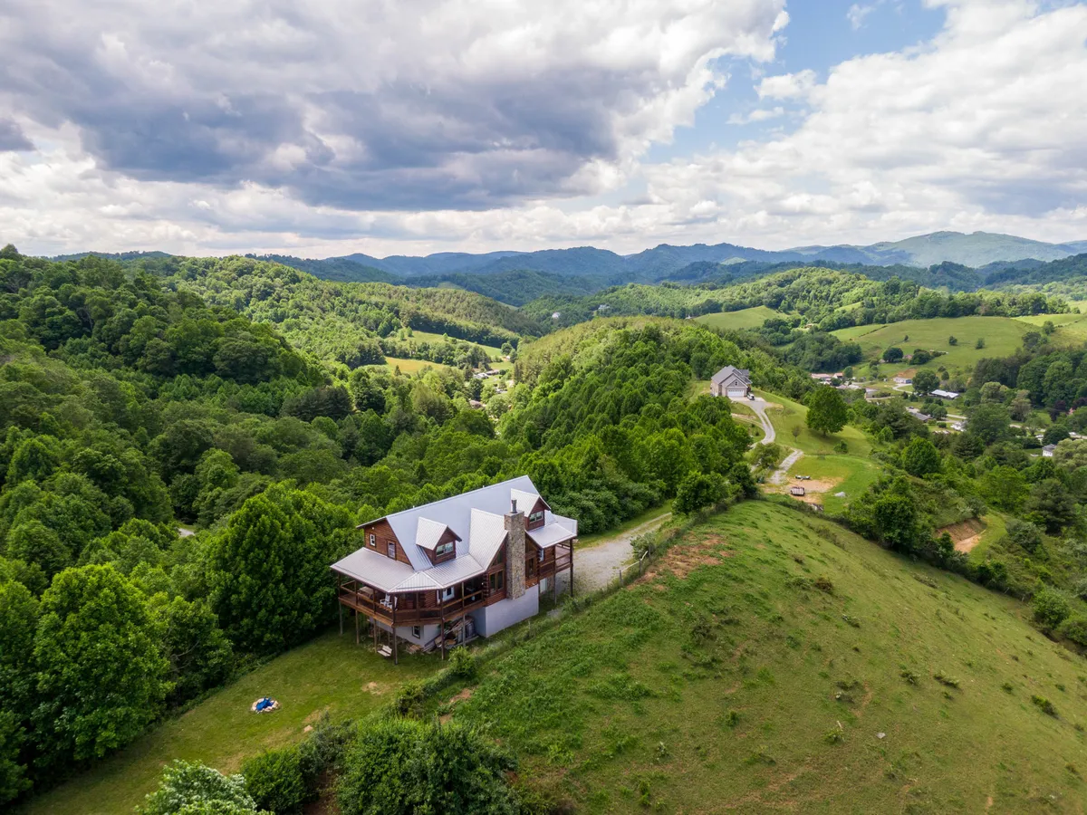 The image showcases a spacious, custom-built mountain home nestled amidst lush greenery, with sweeping views of rolling farmland and a dramatic mountain range along the horizon. The welcoming porch is perfectly positioned to capture sunset vistas, and the tranquil setting hints at opportunities for wildlife spotting and stargazing.