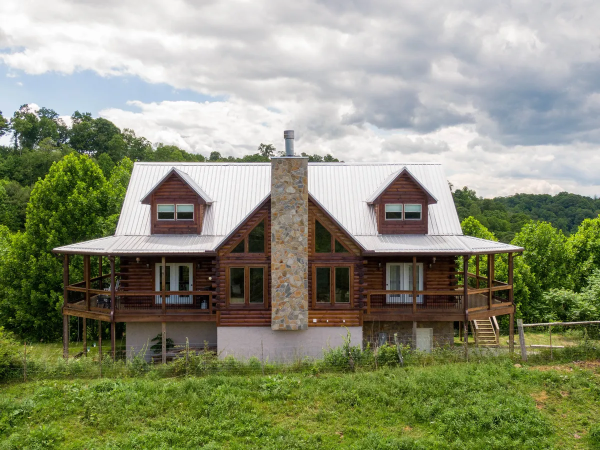 A cozy log cabin structure is framed by a central stone chimney and surrounded by lush greenery. Balconies and a welcoming porch provide inviting outdoor spaces ideal for family gatherings, morning coffee, or unwinding after nature walks. The image conveys warmth, safety, and a pet-friendly atmosphere.