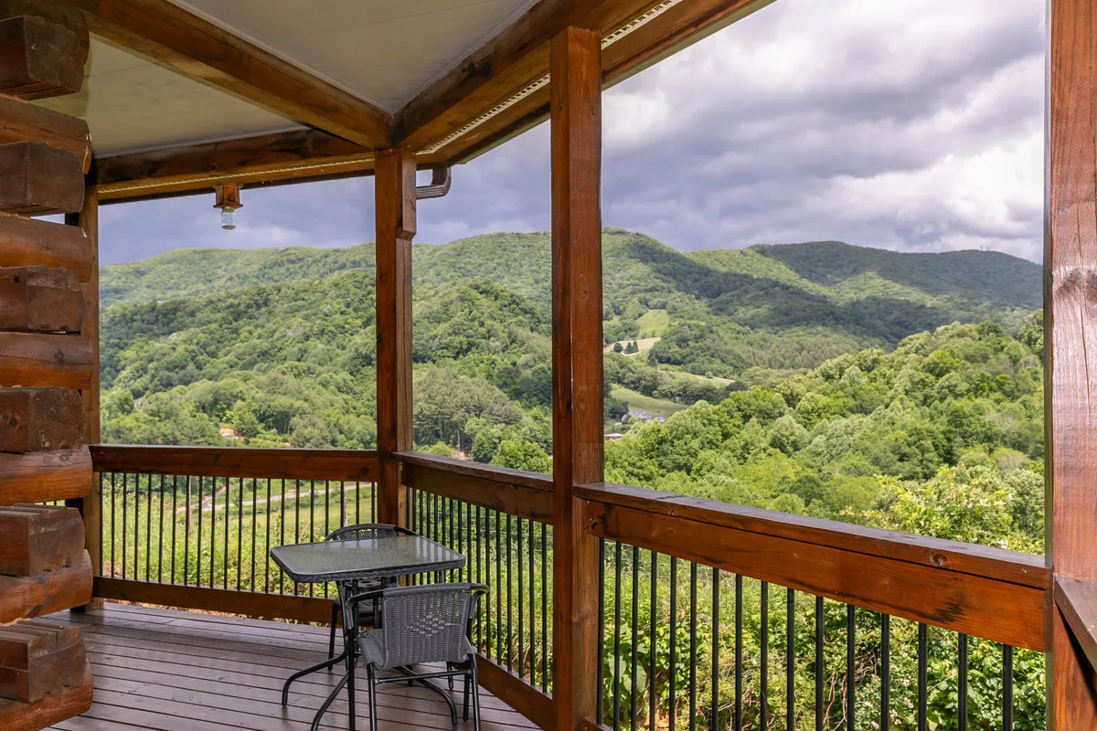 A wooden deck with mountain views, featuring a small table and chairs set along the wooden railing. The scene is tranquil, with soft evening light casting a glow over the rolling farmland and distant peaks. The seating area invites socializing and relaxation, while the open deck connects directly to the outdoors.